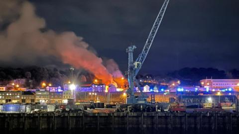 A plumes of smoke come out of a building behind a sea front at night. A crane and boats are in the foreground.