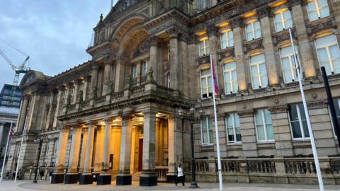 A large and imposing stone building with a columned portico at the front that is lit up, and a domed roof with four flag poles standing in front 