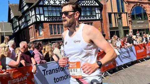 Alex Trivino in a white running vest with an orange number pinned to his chest. He has short dark brown hair, a beard and is wearing dark sunglasses. The man is running in a race. Crowds of people can also be seen in the picture. 