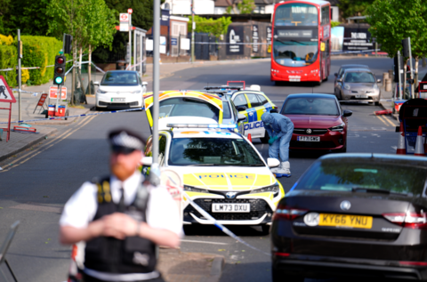 Police cars are parked in the road in the middle of a police cordon around the scene in Golders Green. A scene of crime officer in a blue boiler suit can be seen looking down at the road. A police officer is in the foreground, while an abandoned bus sits in the road in the background.