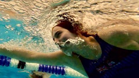 Christina underwater, as she front crawls through the pool. He eyes are open, as she exhales through he nose whilst swimming. She is wearing a blue Team GB swimming costume.