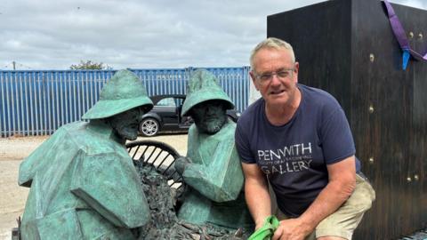 Tom Leaper crouches in front of the artwork he has made. It features two bronze fishermen holding a fishing basket. Tom himself is an older man with short grey hair and glasses, and is wearing a blue T-shirt.