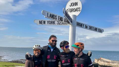 Four men including Ross MacDonald (second left), and Ross MacDonald are standing in front of a white sign which says John O'Groats. The sign has several arms pointing in different directions, one of which says Lands End. The blue sea is in the background and the men are all wearing black.
