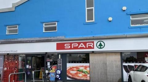 The front of a Spar store is below a tall, blue-painted building.