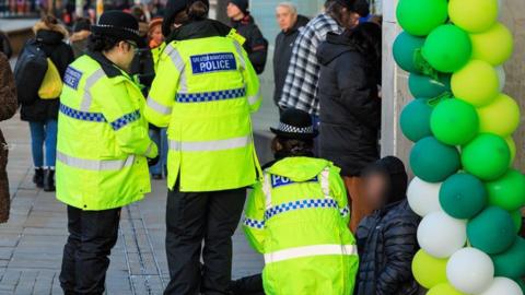 Police officers are talking to a man, whose face is blurred, sitting on a pavement outside some shops. Lime green, green and white balloons are next to him in a doorway.