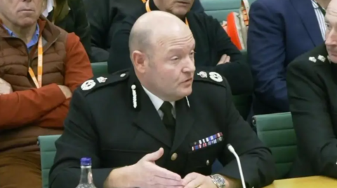 A bald man in a formal police uniform is sitting in a chair with a small microphone in front of him. There are other people around him