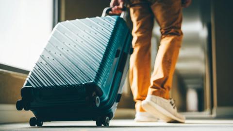 A person wearing beige deck shoes and brown trousers pulls a green, hard cased suitcase along a hotel corridor. Light streams in from a long window.