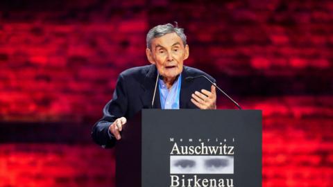 Marian Turski, an elderly man with grey hair wearing a dark blue suit, speaks into small microphones on  a podium which says Memorial Auschwitz Birkenau on it, in front of a brick wall illuminated by red light.