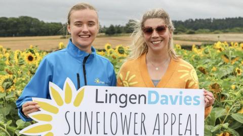 Two women holding a sign which reads Lingen Davies Sunflower Appeal. They are stood in front of a field full of sunflowers. The woman on the left is wearing a blue uniform jacket and the woman on the right is wearing sunglasses and an orange cardigan with stylised images of the sun on it.
