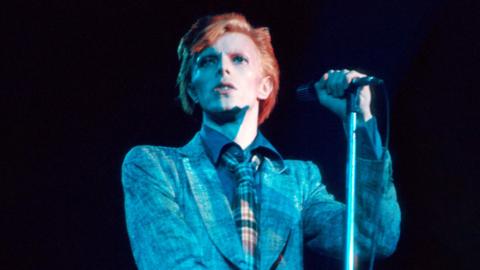 David Bowie performing in 1976, wearing a blue suit, checked tie, red hair, and holding a microphone