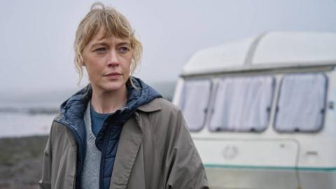 A woman with blonde hair, wearing a grey jacket over a blue hoody. Behind her is a caravan and a seaside view