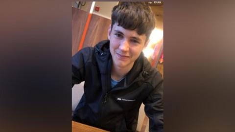 A person wearing a black zip-up jacket sits at a wooden table indoors, leaning slightly toward the camera. The background includes wood-paneled walls and warm indoor lighting. Part of a logo is visible on the jacket, and the image is a close-up portrait-style photograph.