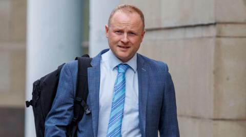 A man with fair, receding hair. He his wearing a blue plaid suit, with a light blue shirt and a tie with different colours of blue stripes. He is carrying a backpack over one shoulder. 