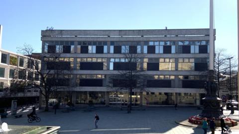 A 1960s style department store has brown tiled panels and large glass windows. It overlooks a war memorial and a pedestrian square