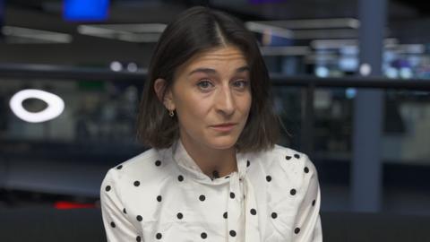 A woman with shoulder length brown hair looks into the camera. She is wearing a predominately white polkadot shirt with black spots. She also has a small golden earring hanging from her right ear.