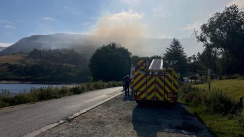 Smoke from a moorland fire rises near the Dove Stone Reservoir. A fire engine is positioned at the front of this image 