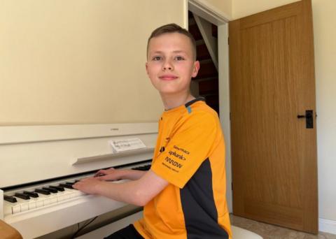 Harvey sits at his white piano and looks at the camera. He wears an orange F1 replica shirt. 