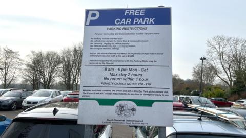 A free parking sign in a car park in Langport in Somerset with multiple parked vehicles in the background. The sign has a list of rules and timings on it