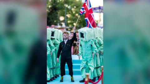 Sam Battle wearing a black jumpsuit holds up the Union Jack flag. People in green suits and square, furry helmets stand behind him.
