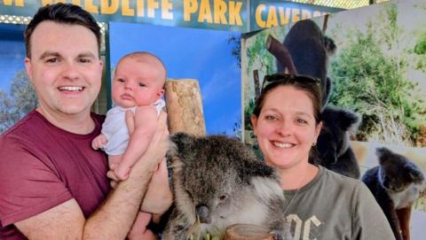 A man carrying a baby, standing with a woman and a koala