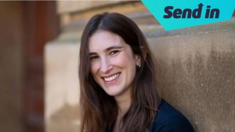 Katherine Rundell smiling leaning against a building. She has long dark hair and is wearing a navy coloured top.