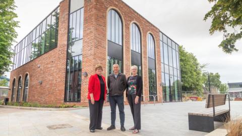 A man flanked by two women pose in front of a new brick and glass building.
