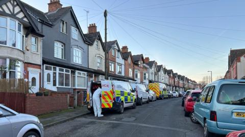 Two police vans on a residential street, there are cars also lined along the street.