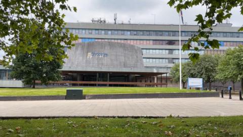 Shropshire Shirehall building which features a large, dark-grey, rounded section, behind which is a six-storey office block. There are trees in the foreground and a large paved area.