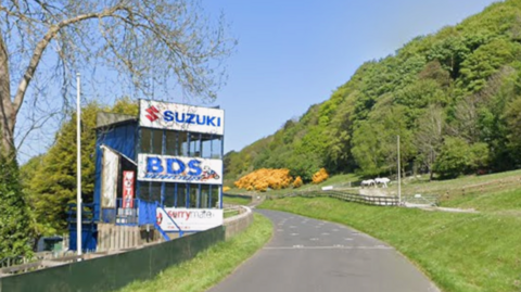 A rural road curves past a blue motorsport viewing stand, with white horses grazing in a fenced field beside green, wooded hills.
