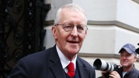 Hilary Benn - he is wearing round glasses and has short white hair. He is wearing a dark suit, dark jumper, white shirt and red tie