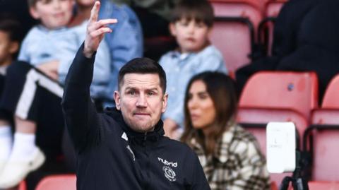 Crewe boss Lee Bell holds his right arm up and shouts instructions from the sidelines during a match.