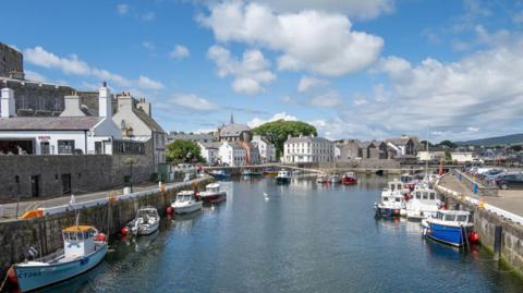 A view of Castletown Harbour on a sunny day. Small boats are moored along both sides of the harbour and there are cars parked on the harbourside on the right. White and grey stone buildings line the area.