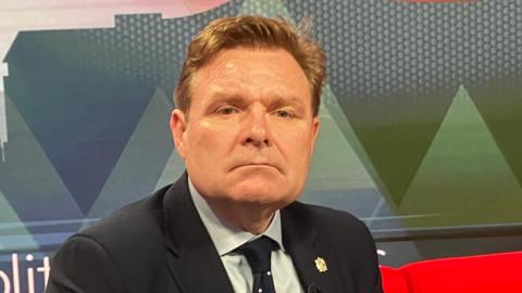 A man with light brown reddish hair sits on a red sofa. He is wearing a black suit jacket, light blue shirt and dark tie. He also has a pin badge on his lapel.