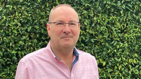 A head and shoulders picture of Bill Mann in his garden with greenery behind him. He is wearing a pink shirt and glasses. 