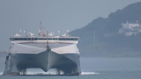 The front view of a DFDS ferry. To the right is land with a house on it. The ferry is black at the bottom and white at the top. It is moving on the water.