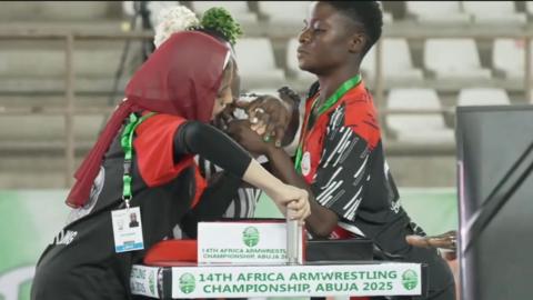 Two female arm wrestlers, one wearing a head scarf, clasp hands and hold handles on a table as a partially-obscured referee prepares them for a contest