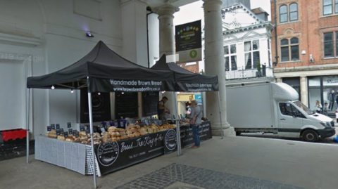 The image shows two black stalls with black awning and white writing that says "Handmade Brown Bread". Stacked loaves of bread can be seen on the stalls and a man can be seen selling some to a woman.