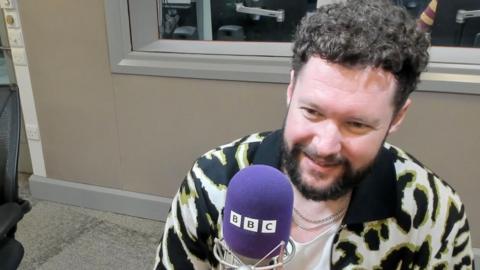 Calum Scott sits in a radio studio setting behind a purple mic with the letters BBC