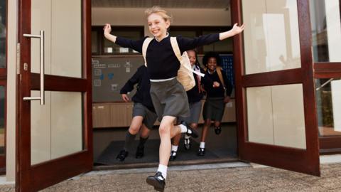 Girl runs out of the school entrance with her arms in the air and a big smile on her face. She is wearing her school uniform and other pupils are following her