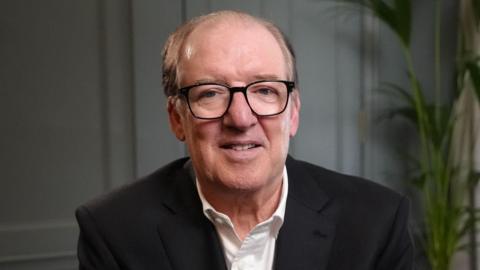 David Murray looking at the camera. He is balding with black glasses and is wearing a white shirt and dark jacket 