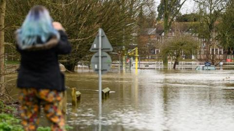 A woman can be seen from behind holding a phone and taking pictures of submerged cars in a car park after heavy rains and sewer system overflows caused the River Thames to break its banks, on 5 January 2024. She is wearing a blue fleece and floral trousers - she also has died blue hair.