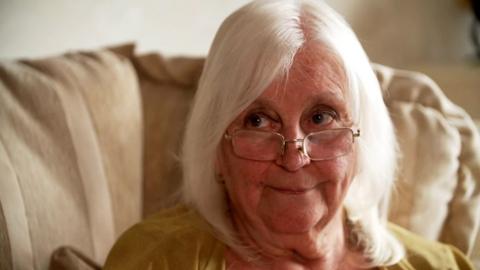 Sue is sitting on her cream-coloured sofa. She has white shoulder length hair and a pair of glasses perched on her nose. She is wearing a mustard top.