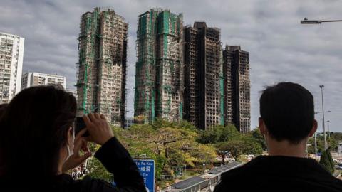The back of two people's heads as they look at four charred high-rise blocks in the distance, with trees and roads between them and the towers