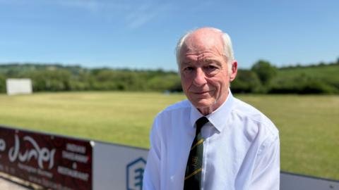 Tony Evans, a man with white hair, sits with his back to the cricket match, wearing a white shirt and green and yellow Purnell Cricket Club tie.