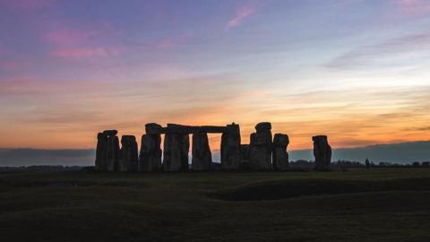 A sunset at Stonehenge, with the stones obscured in darkness and an orange, pink and blue coloured sky in the background