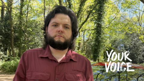  A dark-haired bearded man in a red shirt standing in front of some trees 