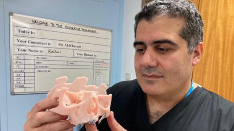 A man wearing a black surgeon's scrubs holding a pink 3D resin print out of the top half of a skull 