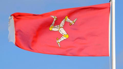 In front of a bright blue sky, the Isle of Man flag flies on a flagpole. The flag is red, in the middle is a triskelion made of three interlocked legs bent at the knee clad in metallic armour. 