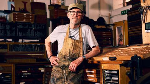 Nick Hand wearing a grey t-shirt and brown apron, which is heavily stained with black ink. He is leaning on a wooden table inside his workshop, looking straight at the camera with his hand on his hip.