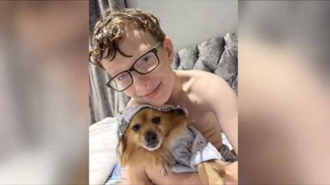 An 11-year-old boy wearing glasses with brown hair holding a brown dog to his chest
